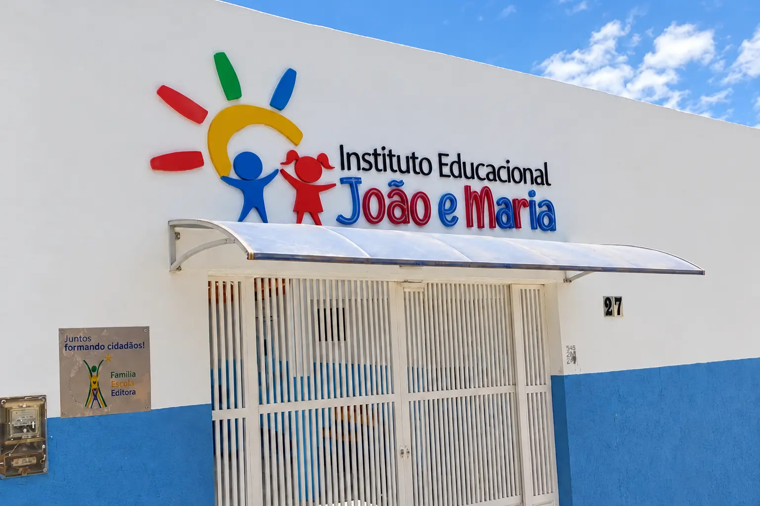Fachada do Instituto Educacional João e Maria, com letreiro, portão branco e faixa azul