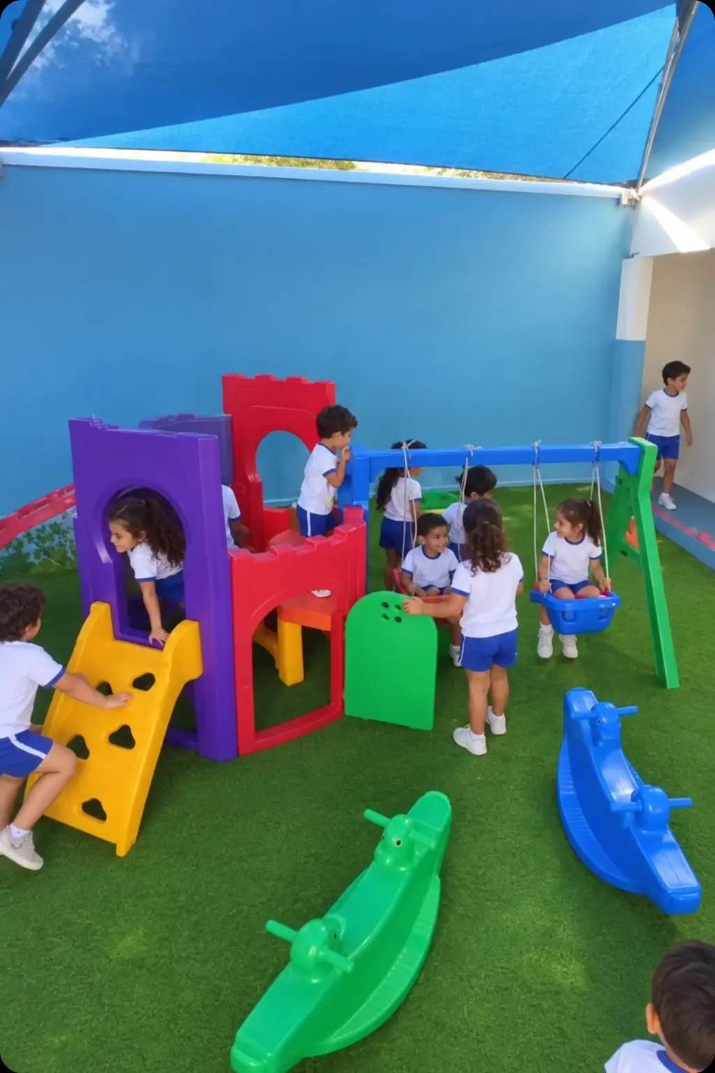 Alunos de farda branca e short azul brincando no parquinho coberto, com grama sintética, do Instituto João e Maria